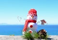 Bright red and white handmade snowman soft toy fleece snowman by bright blue sea horizon background with pine cone & pine needles Royalty Free Stock Photo