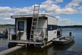 White square shaped house boat with ladder leaning on front wall and wooden bridge in front