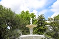 A white stone status on the tops of a water fountain surrounded by lush green trees and plants with blue sky and clouds Royalty Free Stock Photo