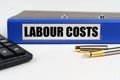 On a white surface, a calculator, a pen and a folder with the inscription - LABOUR COSTS