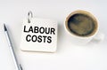On a white surface, a cup of coffee, a pen and a notepad with the inscription - LABOUR COSTS