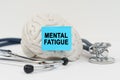 On a white surface next to the stethoscope lies a brain on which a sticker with the inscription - Mental Fatigue