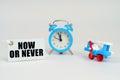 On a white surface there is a toy plane, an alarm clock and a sign with the inscription - Now or Never