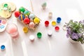 On a white table, in daylight, there are brushes in a glass of water, multi-colored eggs in a tray, jars of paint.