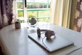 On a white table there is a metal white desktop fan, a flower in a flowerpot, a closed laptop, wired headphones are on the laptop,