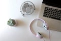 On a white table there is a metal white desktop fan, a flower in a flowerpot, a closed laptop, wired headphones are on the laptop,