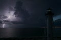 White tall lighthouse on sea shore with blinking light at stormy night for commercial vessels navigation. Thunderstorm