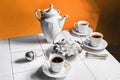 White tea cups and teapot, bowl with sugar cubes, tongs and tea stainer on white tiles table over orange background