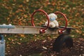 white teddy bear children soft toy with disposal face mask sits lonely on a wooden seesaw of an outdoor playground during autumn.