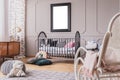 White teddy bear on the floor of stylish baby room interior with grey wooden crib with pillows, white rocking chair and mockup