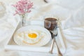 White tray with breakfast on a bed in a hotel room. Fried egg, cup of coffee and flowers in white sheets in light bedroom Royalty Free Stock Photo
