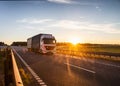 A white truck with a semitrailer carries cargo along the highway against the backdrop of a sunny sunset in the evening