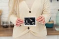 White unrecognisable woman wearing a knitted sweater holding up ultrasound photo of baby to the camera. Close up Indoor