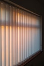White vertical slat blinds hanging in front of a window as the sun is setting turning the light golden. Royalty Free Stock Photo