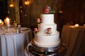 White wedding cake with red roses on a silver platter with a wedding table and candles in the background Royalty Free Stock Photo