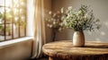 White wildflowers in paunchy vase on round old wooden brown table against empty gray wall. Natural side lighting from window