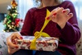 Blonde woman unwraps a Christmas present with the Christmas tree in the background with the lights on Royalty Free Stock Photo