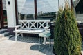 A white wood table and bench stand unoccupied on a sunny summer day against a glass wall. Coffee shop terrace. Cozy cafe. A place