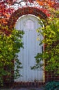 White wooden arched garden gate in autumn in brick wall surrounded by foliage with the sunlight shining around it and through colo Royalty Free Stock Photo