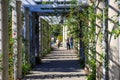 White wooden awning in the garden covered with lush green plants or colorful flowers with stone pillars and people walking