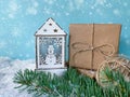 White wooden candle lantern with snowman next to handcrafted gift and twine ball over snowy fir tree branch.