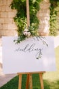 White wooden easel with an inscription on the background of a stone wall and greenery. Caption: Welcome to our wedding