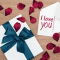 White wrapped gift with a turquoise ribbon and a I-love-you-note with a white envelope on a wooden board, surrounded by red rose p Royalty Free Stock Photo