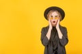 Young cheeky blonde woman in a black suit, hat and glasses is surprised on a yellow background