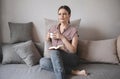 1 white young woman in pink blouse and gray jeans sits on couch with mug in the hands of and with easy smile looks in side,