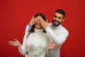 Who is it. Positive arab man covering wife& x27;s eyes from back, standing behind her and smiling, red background