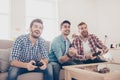 Who will win? Competition of guys playing car race. Excited friends are playing games indoors at home, sitting on cozy beige Royalty Free Stock Photo