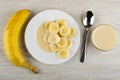 Whole banana, white plate with slices of banana and condensed milk, spoon, bowl with milk on wooden table. Top view Royalty Free Stock Photo