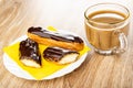 Whole eclair and halves of eclair with chocolate on yellow napkin in plate, cup of coffee with milk on wooden table Royalty Free Stock Photo