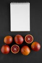 Whole and halved blood oranges, blank notebook on black background, top view. Flat lay, overhead, from above Royalty Free Stock Photo