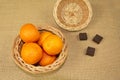 A wicker basket with a lid, tangerines, three pieces of chocolate on a table covered with burlap.