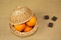 A wicker basket with tangerines, covered with a lid, three pieces of chocolate on a table covered with burlap.