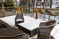 Wicker garden furniture on a wooden, snowy terrace with glass balustrades, in the background trees and houses.