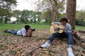 Wide angle shot of photographer taking a photo of relaxed young man with headphones playing acoustic guitar and sitting under a tr