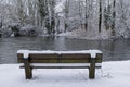 Wide angle shot of a wooden bench in front of the water in a park covered in snow
