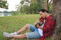 Wide angle shot of young relaxed man in red shirt leaning against a tree and playing acoustic guitar in beautiful outdoor park. Royalty Free Stock Photo