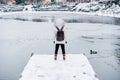 Wide angle view of back view of backpacker woman standing in front of frozen lake in city. Woman on pier. Lifestyle during winter