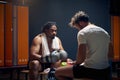 Wide horizontal shot of strong muscular boxing player after workout in sweats resting on bench with coach