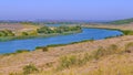 A wide river flowing between fields and low slopes covered with dry grass with peacefully grazing cows in the meadow. Royalty Free Stock Photo