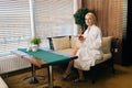 Wide shot of beautiful blonde woman in white bathrobe holding cup with fresh herbal tea smiling looking at camera Royalty Free Stock Photo