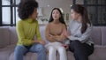 Wide shot portrait of sad brokenhearted beautiful Asian woman sitting on couch as African American and Caucasian women