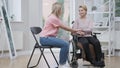 Wide shot of sad disabled young woman in wheelchair and supportive friend holding hands. Blond Caucasian empathic lady