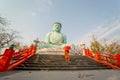 Wide shot side view back of young Asian monk hold red umbrella and walk up on stair in front of green big buddha statue with soft