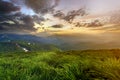 Wide summer mountain view at sunrise. Glowing orange sun raising in blue cloudy sky over green grassy hill soft grass and distant Royalty Free Stock Photo