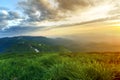 Wide summer mountain view at sunrise. Glowing orange sun raising in blue cloudy sky over green grassy hill soft grass and distant Royalty Free Stock Photo