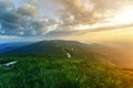 Wide summer mountain view at sunrise. Glowing orange sun raising in blue cloudy sky over green grassy hill soft grass and distant Royalty Free Stock Photo